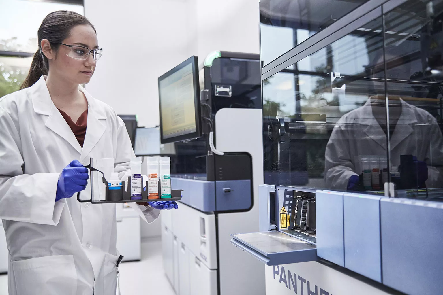 Vrouwelijke arts houdt een tray met buisjes vast naast het Panther systeem in een laboratoriumomgeving.